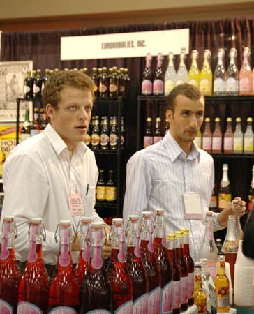 Two men in white shirts and name badges stand behind rows of red and pink bottled drinks at a booth with a Eurobubbles, Inc. sign and shelves of bottles behind them.