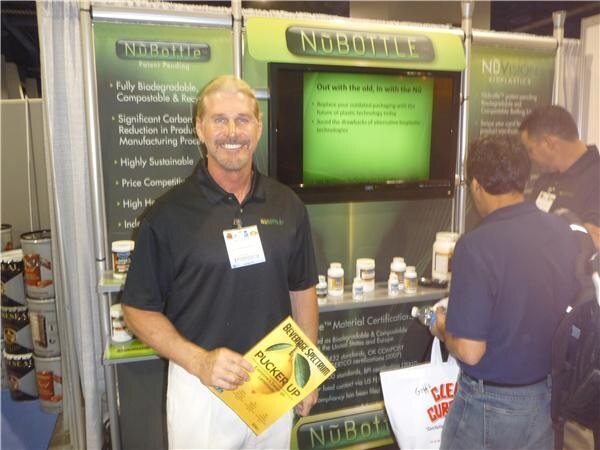 A smiling man in a black polo holds a yellow magazine in the NuBottle booth, which has a screen and display of bottles. Two other people stand in the background.