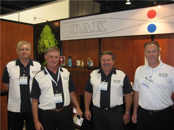 Four men with name badges stand side by side in front of a wood-paneled Oak booth with an OAK sign. Three wear black and white bowling-style shirts and one wears a white polo.