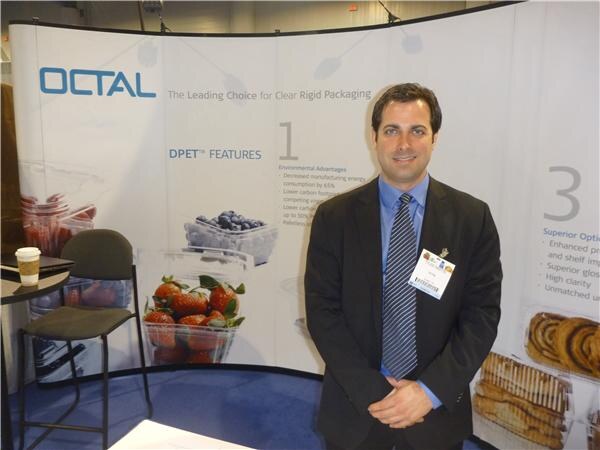 A man in a dark suit and blue shirt with a name badge stands in front of an Octal booth display about clear rigid packaging, with images of strawberries and blueberries in clear containers.