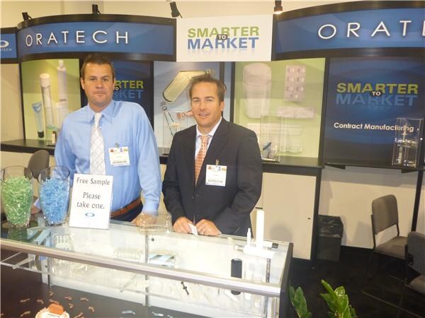 Two men with name badges stand behind a counter at the Oratech booth under Smarter to Market signs. A Free Sample sign and bowls of colored pellets sit on the counter.