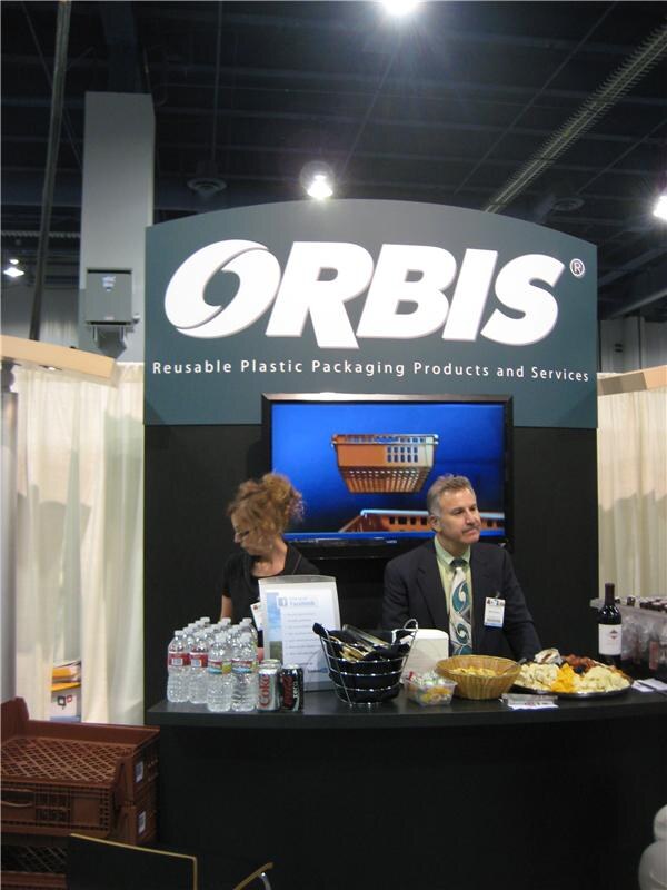 A man in a dark suit and a woman with upswept hair stand behind the counter of the Orbis booth. Water bottles, snacks and a stack of plastic crates are visible, with a screen showing a basket.