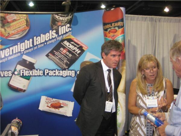 A man in a dark suit and a woman with glasses talk at the Overnight Labels, Inc. booth, with another person at the right edge holding a can. Package samples are on the display wall.