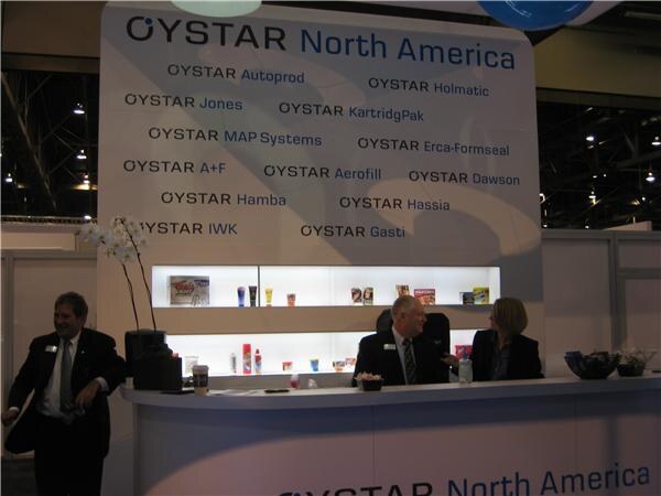 The OYSTAR North America booth has a wall listing OYSTAR brand names. A man stands at the left, and a man and a woman sit behind the counter beside a lit shelf of packaged products.