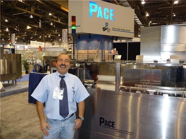 A smiling man with a mustache and a name badge stands in front of a stainless steel Pace machine. A Pace sign hangs overhead, and an OYSTAR sign is visible in the background.
