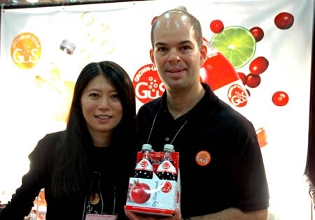 A woman and a man in black shirts stand together, the man holding a four-pack of GuS cranberry soda bottles, in front of a GuS backdrop with fruit graphics.