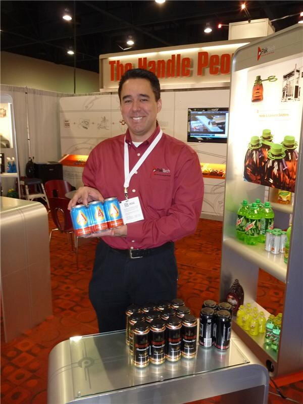 A smiling man in a red PakTech shirt holds three blue cans joined by a plastic handle. Cans and bottles sit on a table in front of him, and a shelf of bottles is at the right.