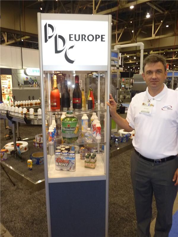 A man in a white polo shirt and name badge stands beside a lit display case labeled PDC Europe. It holds sleeved bottles and cans, including Heineken and Coors Light packages.