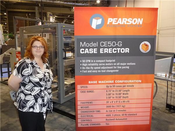 A woman with red hair and glasses, in a black and white patterned top, stands beside a Pearson Packaging Systems banner for the Model CE50-G case erector, with a machine behind her.