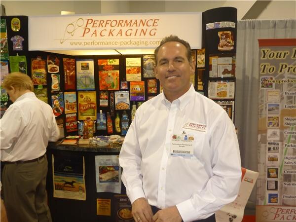 A smiling man in a white button-up shirt with a name badge stands at the Performance Packaging booth, where sample pouches and packages are displayed on the back wall. Another man stands at left.