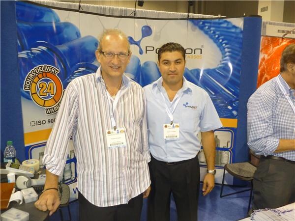 Two men wearing convention badges pose side by side in front of a blue Polykon backdrop showing plastic containers and a 24-hour delivery graphic. A third man stands partly visible at right.