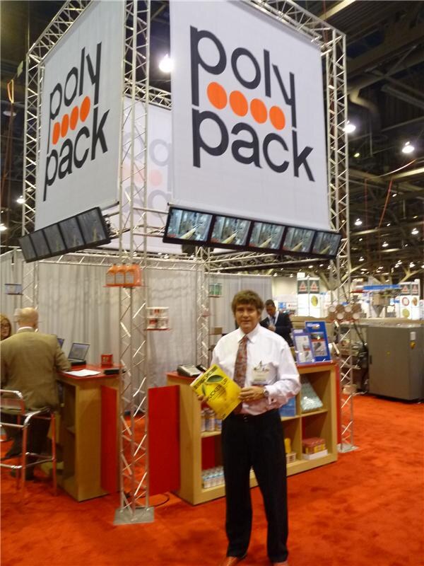 A man in a white shirt and tie holds a yellow booklet beneath large hanging poly pack banners in a truss-framed booth with wooden display shelves on an orange carpet. A seated man works at a counter.