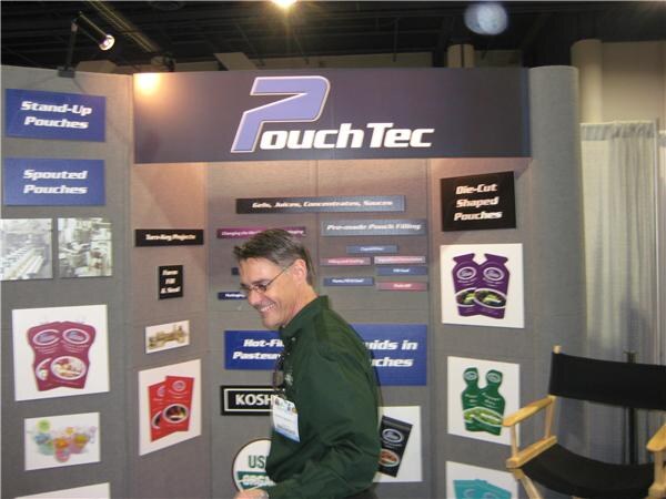 A man with glasses in a dark green shirt walks past the PouchTec booth, which has panels about stand-up and spouted pouches and displays of printed pouches. A folding director's chair sits at right.