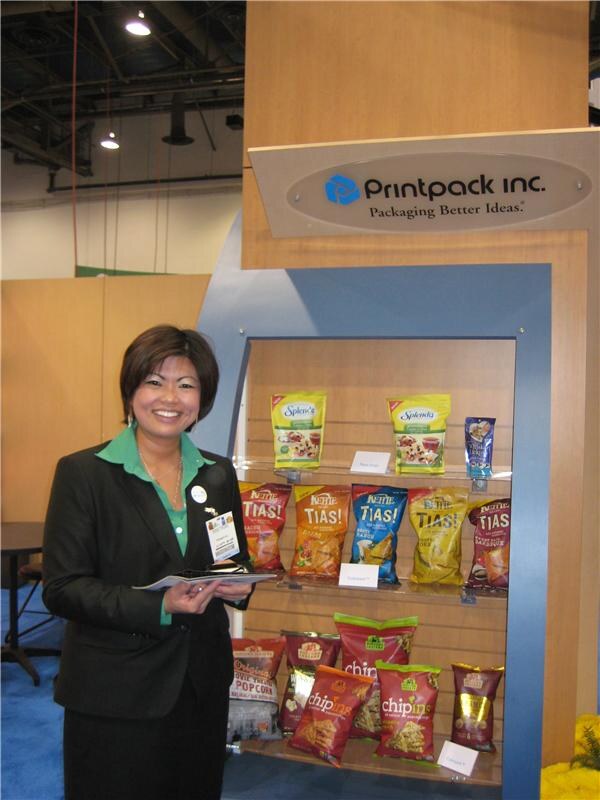 A smiling woman in a dark blazer and green blouse holds a folder beside a Printpack Inc. display shelf of snack bags, including Splenda, Nettie Tias! and Chip-ins packages.