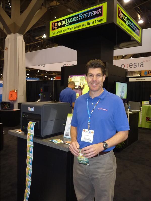 A smiling man in a blue polo shirt with a lanyard stands beside a label printer with a strip of printed color labels at the QuickLabel Systems booth. A Coesia sign hangs in the background.