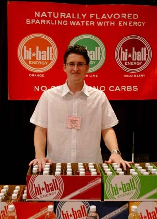 A man with glasses and a white shirt stands behind cases of hi-ball Energy sparkling water, beneath a red banner reading "Naturally flavored sparkling water with energy".