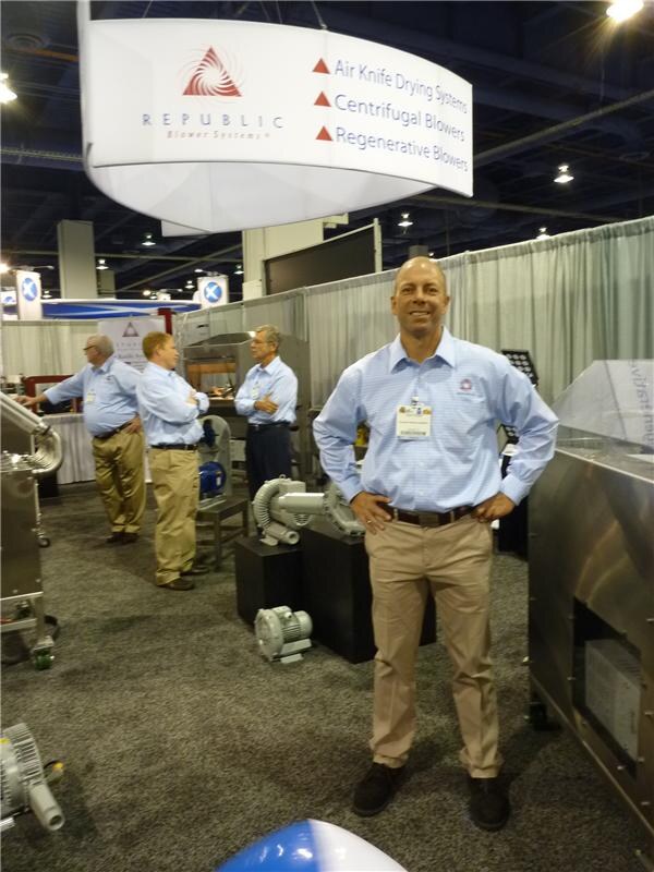 A man in a light blue shirt and khaki pants stands with hands on hips under a Republic Blower Systems sign listing air knife drying systems and blowers. Three men talk behind him near blower equipment.