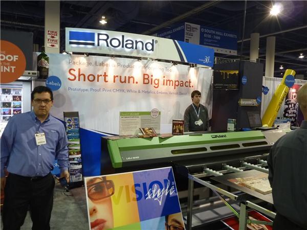 A man in a blue shirt with a badge stands at left in the Roland booth, where a wide-format printer sits under a banner reading Short run. Big impact. Another man stands behind the printer.