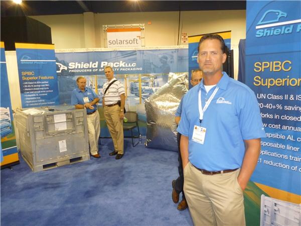 A man in a blue polo shirt and lanyard stands in the foreground at the Shield Pack booth. Behind him, two men stand near a collapsible container and a Solarsoft sign, with SPIBC banners at the sides.