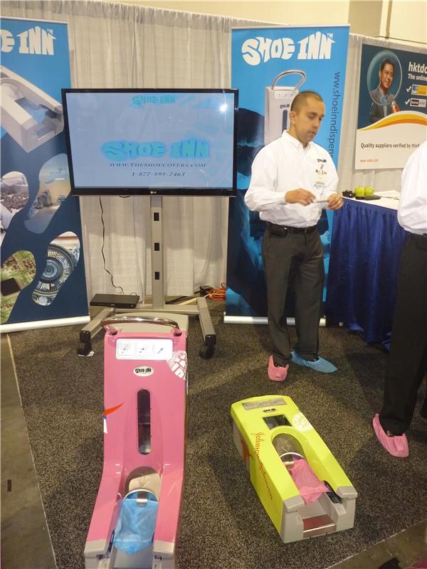 Shoe Inn booth with an LG screen showing the Shoe Inn logo, a man in a white shirt and shoe covers, and a pink and a yellow-green shoe cover machine on the carpeted floor.