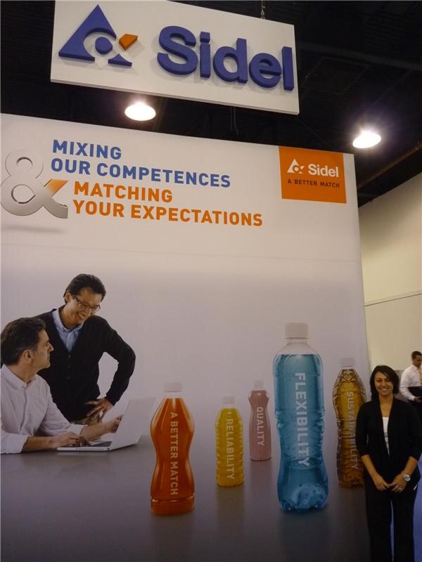 Sidel booth with a large backdrop reading Mixing our competences & matching your expectations, oversized colorful bottles, and a smiling woman in black standing at the right.