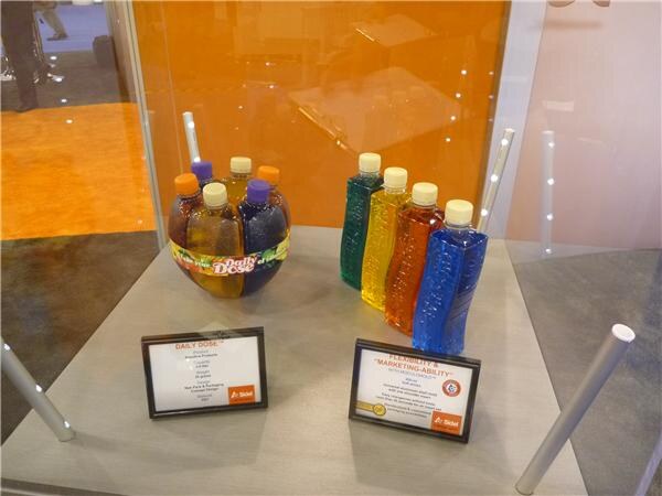 Display case with a round cluster of small bottles labeled Daily Dose on the left and a row of tall colored bottles on the right, each with a Sidel information card in front.