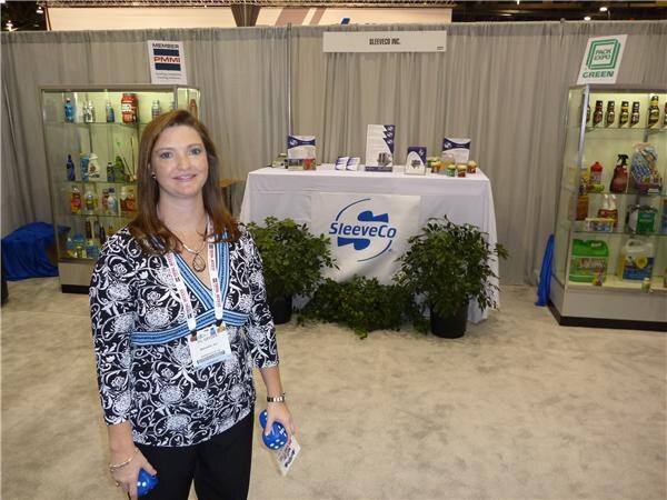 A smiling woman in a black and white patterned top with a badge stands in front of the SleeveCo booth, which has a table with a SleeveCo banner, plants and product display cases.
