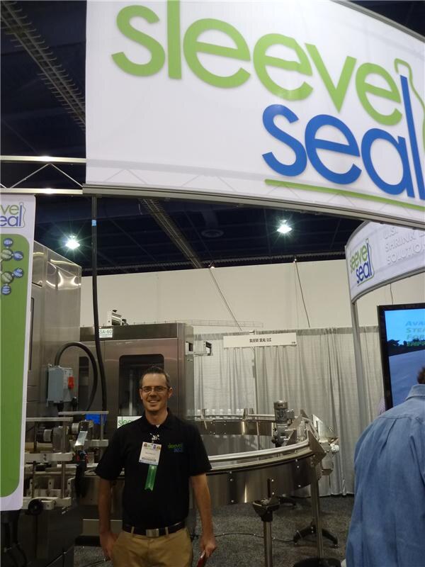 A smiling man in a black polo and glasses with a show badge stands in the Sleeve Seal booth, under a large Sleeve Seal sign, with labeling machinery behind him.