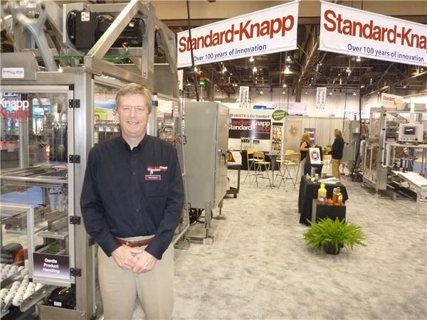 A smiling man in a dark shirt and khakis stands beside packaging equipment, with Standard-Knapp banners hanging above the booth behind him.