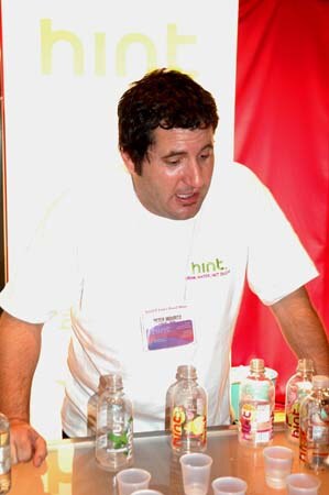 A dark-haired man in a white hint T-shirt leans over a table of bottled hint flavored waters and small plastic sample cups, in front of a hint banner.