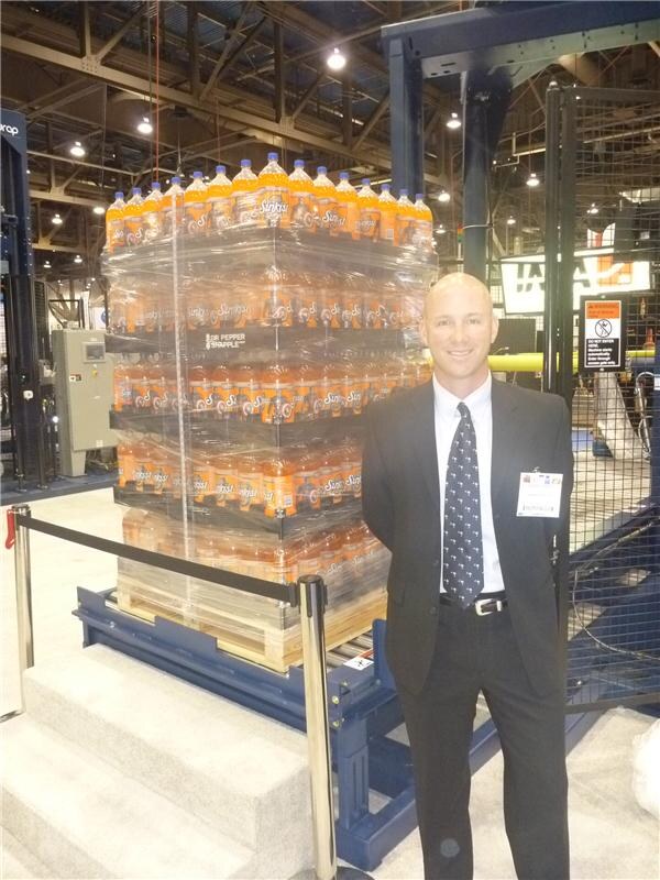 A smiling bald man in a dark suit and badge stands beside a shrink-wrapped pallet of orange Sunkist soda bottles marked Dr Pepper Snapple, next to a blue conveyor and safety fencing.