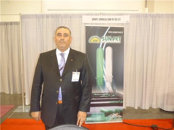 A man in a dark suit and tie with a badge stands beside a Sun Pet banner showing green and white plastic preforms, in front of white draped booth walls.
