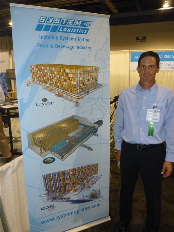 A man in a light blue shirt with a badge stands beside a System Logistics banner showing automated warehouse illustrations and the text Installed Systems in the Food & Beverage Industry.