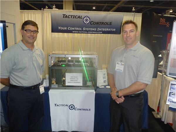 Two men in gray polo shirts and badges stand on either side of a clear display case under a Tactical Controls banner reading 'Your Control Systems Integrator'.