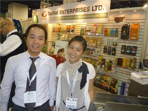 A smiling man in a striped shirt and tie and a smiling woman in a polka-dot top, both wearing badges, stand before a Taipak Enterprises Ltd. wall of packaging pouches.