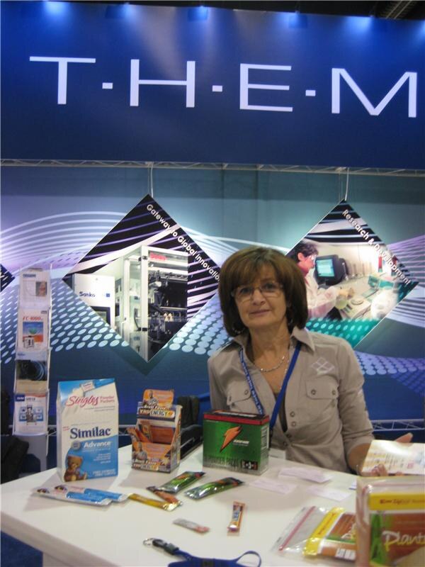 A woman with glasses sits at a white table in the T.H.E.M. booth, with packaged samples including Similac and Gatorade items on the table and a T.H.E.M. sign behind her.