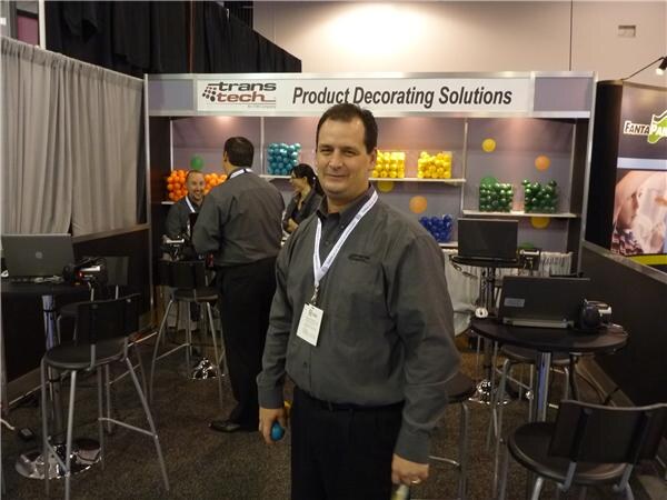 A man in a gray button-up shirt and lanyard stands in front of a Trans Tech booth reading 'Product Decorating Solutions', with colored balls on shelves and other staff behind him.