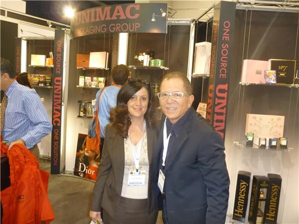A woman and a man wearing badges pose at the Unimac Packaging Group booth, with shelves of gift boxes and Hennessy and Dior packaging displayed behind them.