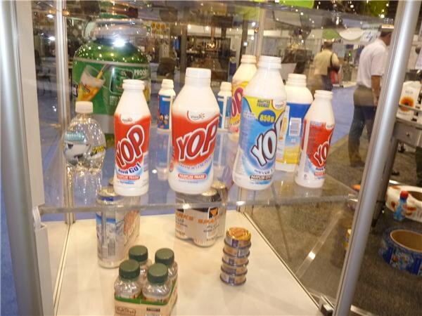 Inside a glass display case are several white YOP bottles with red and blue sleeves, plus small water bottles and cans. Visitors can be seen in the aisle behind the glass.