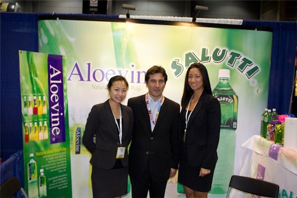 A man in a dark suit stands between two women in dark blazers in front of Aloevine and Salutti banners, with green bottles of aloe drink on a table at right.
