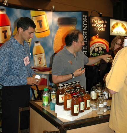 Two men at the Honest Tea booth; one in a blue patterned shirt holds a small cup, the other in a gray polo reaches over a table of tea bottles. A woman stands behind them.