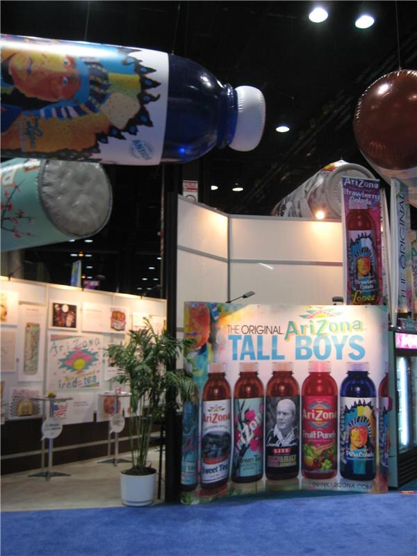An AriZona booth with oversized inflatable bottle and can displays overhead and a 'The Original AriZona Tall Boys' sign showing a row of AriZona tall cans.