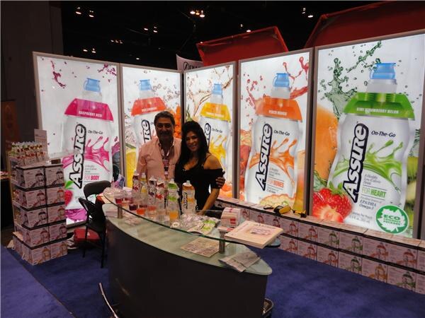 A man and a woman stand behind a curved glass counter at the Assure booth, with large backlit panels of Assure On-The-Go bottles and fruit behind them and product samples on the counter.