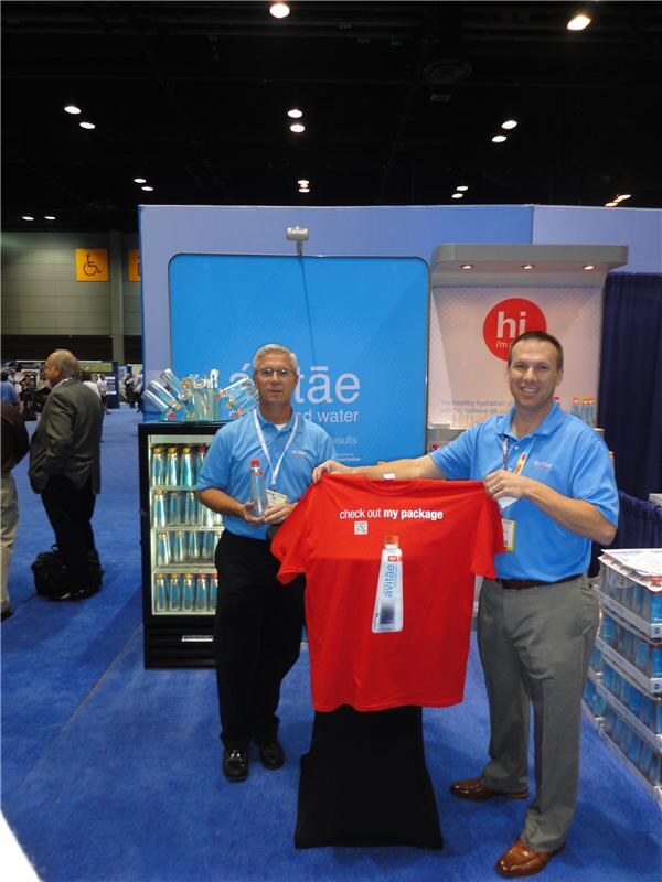 Two men in blue polo shirts and lanyards hold up a red t-shirt reading 'check out my package' with an avitäe bottle on it, in front of the avitäe booth and a cooler of bottles.