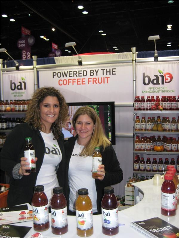 Two smiling women in white bai t-shirts hold bottles of bai drinks at a booth with a 'Powered by the Coffee Fruit' sign, bai5 shelves of bottles behind them and more bottles on the counter.