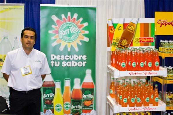 A man in a white shirt stands beside a green Barrilitos banner reading Descubre tu sabor. Shelves of orange Topo Sabores bottles stand at right.