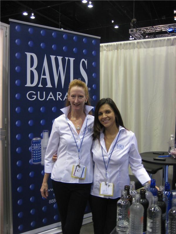 Two women in white BAWLS button-up shirts and lanyards stand side by side in front of a blue BAWLS banner, with clear and dark bottles on a table in the foreground.