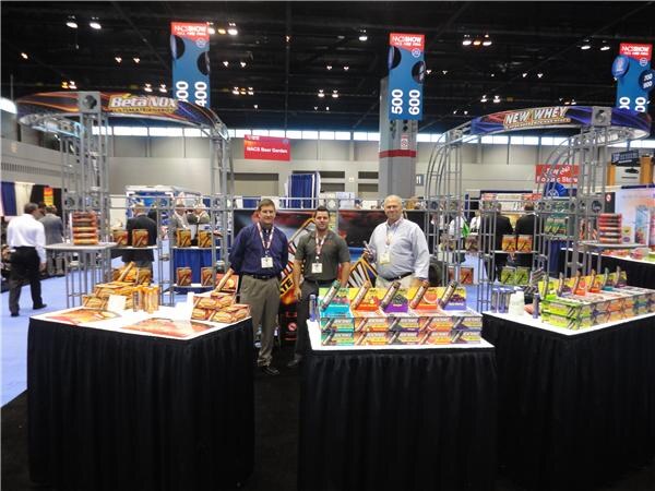 Three men stand between two display tables at a booth with truss arches labeled Beta NOX and New Whey. Product boxes are stacked on the tables, which are draped in black cloth.