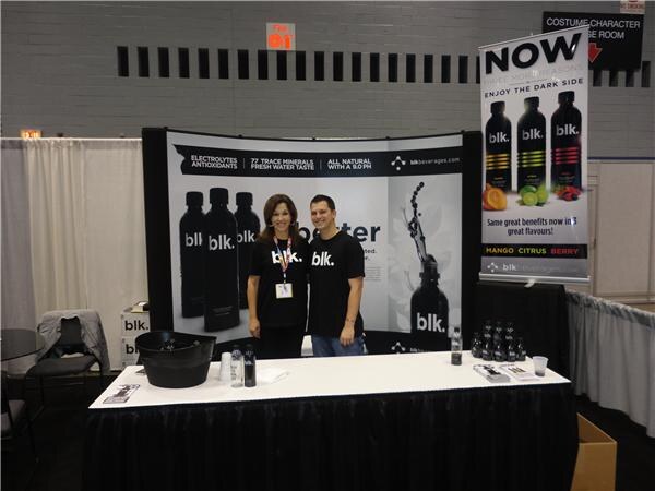 A woman and a man in black blk. t-shirts stand behind a draped table of blk. water bottles, with a backdrop of black bottles and a banner listing mango, citrus and berry flavors.