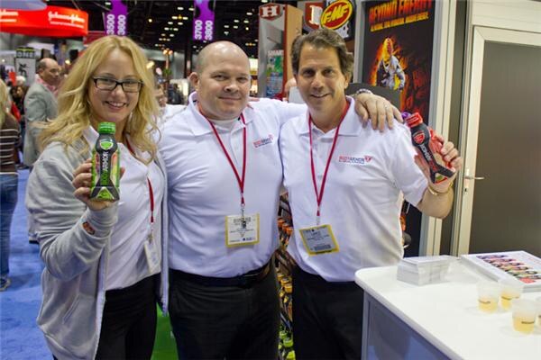 A woman holding a green bottle and two men, one holding a red bottle, pose with their arms around each other in white BodyArmor polo shirts and lanyards at a booth with sample cups.